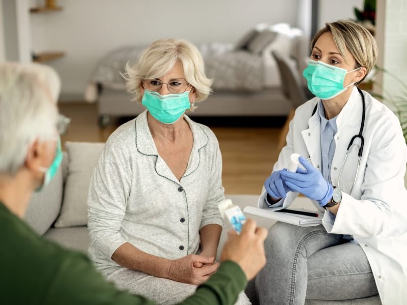 Female doctor talking to senior couple while vising them at home during coronavirus epidemic. They are wearing protective face masks.