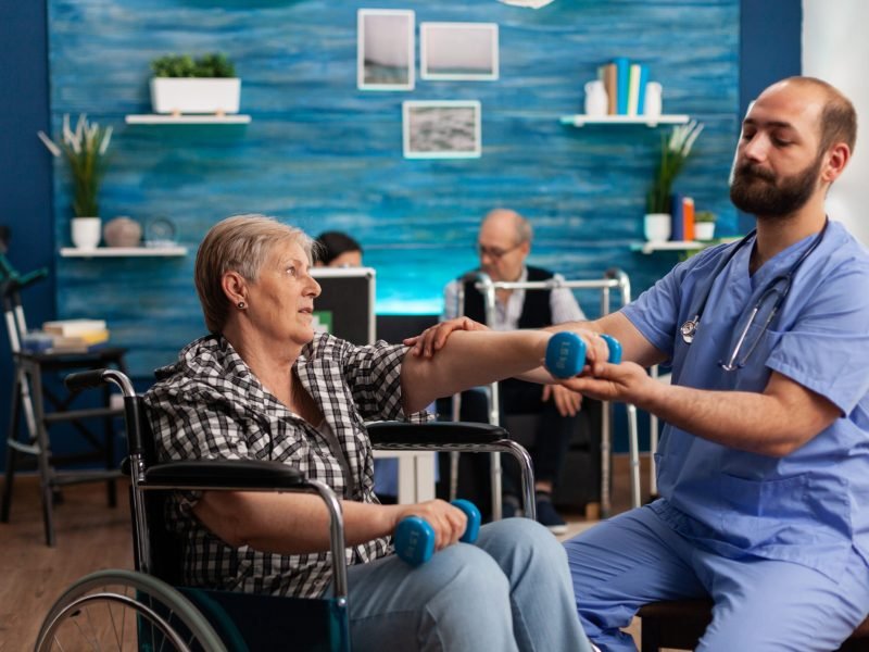 Male nurse helping senior retired disabled woman in wheelchair to rehabilitate using dumbbels during recovery session in nursing home. Elderly woman exercising physiotherapy with social worker