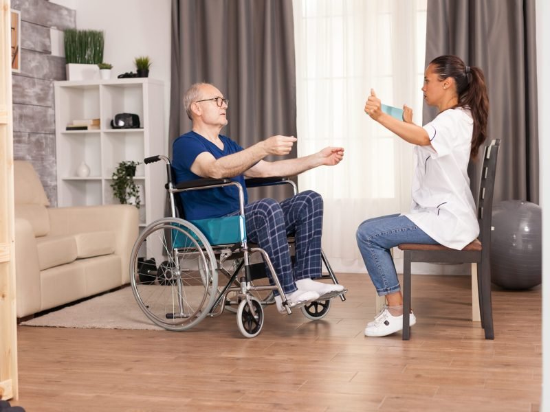 Nurse using resistance band and explain to the old man how to use it. Disabled handicapped old person with social worker in recovery support therapy physiotherapy healthcare system nursing retirement home